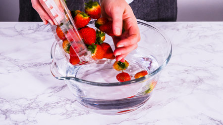 Ripe strawberries are submerged in water within a large glass mixing bowl, a step in washing the fruit to ensure cleanliness and longevity before storage or consumption.の写真素材