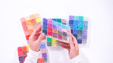Flat lay. Woman hands gracefully poised over a collection of beads, sorted by color in transparent...