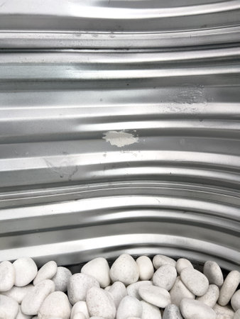 A close-up view of a corrugated metal window well with white decorative rocks at the base. The metal surface has minor scratches and wear, reflecting natural light. This functional design is commonly used for basement windows to allow light and ventilation while preventing soil erosion and water buildup.の写真素材