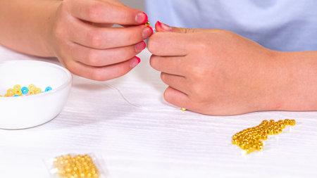 Delicate fingers of a young girl navigate through a treasure trove of bright, multicolored beads, each compartment revealing a new hue to choose from. She immersed in the joyful task of stringing together a handmade bracelet, with golden beads and pearls lying nearby to add a touch of sparkle to her creation.の写真素材