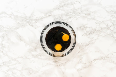A top-down view of a glass bowl containing two cracked eggs and dark molasses, key wet ingredients in the Zesty Orange Gingerbread Cookie Dough recipe. The yolks contrast with the rich molasses, set against a marble background.の写真素材