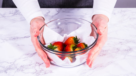 Strawberries, freshly washed and dried, are neatly stored in a glass bowl lined with a paper towel and securely covered with plastic wrap to maintain freshness.の写真素材
