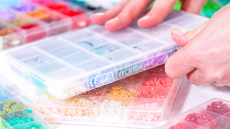 Woman hands gracefully poised over a collection of beads, sorted by color in transparent organizers....
