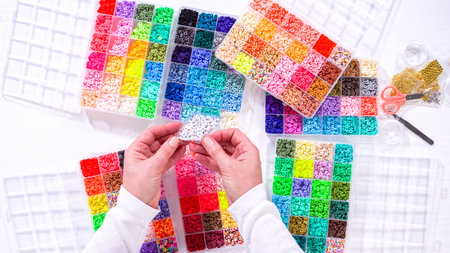 Flat lay. Woman hands gracefully poised over a collection of beads, sorted by color in transparent...
