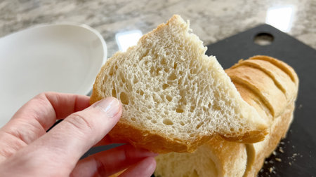 Close-up of a hand holding a slice of white bread in front of a loaf resting on a black cutting...
