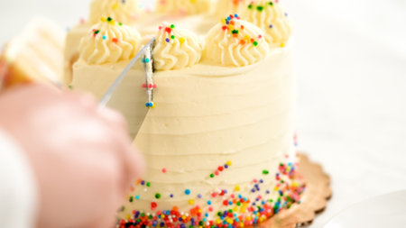 Close-up of a hand using a knife to slice into a cake with piped frosting and colorful sprinkles. The cake sits on a gold base over a marble surface, ready to serve.の写真素材