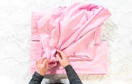 A person folds a soft pink bed sheet on a white marble surface. Hands are visible organizing the sheet, highlighting a clean and minimal setting with natural light.の写真素材