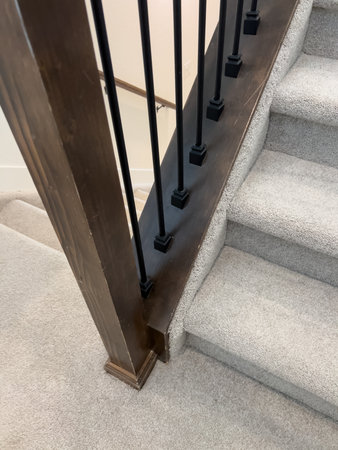 Close-up of the base of a staircase with a wood post, black spindles, and beige carpeted steps. The image highlights the structural transition between materials in a modern home.の写真素材