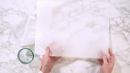 Two hands lift a full-sized sheet of parchment paper over a kitchen counter. A glass and folded paper cone sit nearby.の写真素材