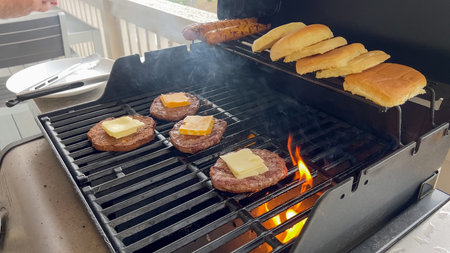 Four beef patties with melting slices of cheese cook on a gas grill beside hot dog sausages and sandwich buns. The grill flames are visible beneath the patties, adding a smoky, authentic cookout feel. Perfect for summer BBQ themes, backyard gatherings, or food advertising.の写真素材