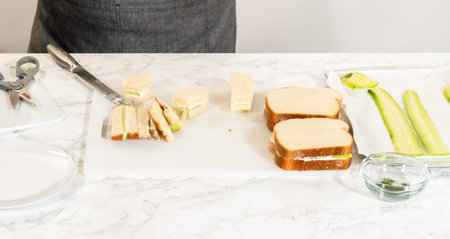 Using a serrated knife, cucumber sandwiches are being sliced on a prep surface. Kitchen scissors,...