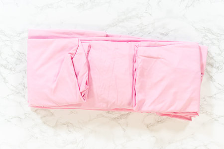A light pink bedsheet begins to unfold across a white marble background. The fabric is soft and...