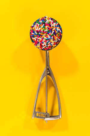 A metal cookie scoop filled with colorful rainbow sprinkles is placed on a bright yellow background....