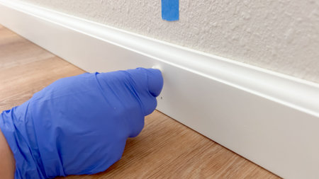 Close-up of a hand in a blue glove inspecting or cleaning a white baseboard trim set against a tan...