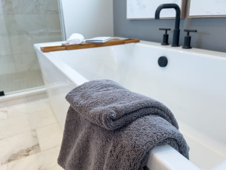 A white modern soaking bathtub with a folded gray towel draped over the side. The tub features matte...