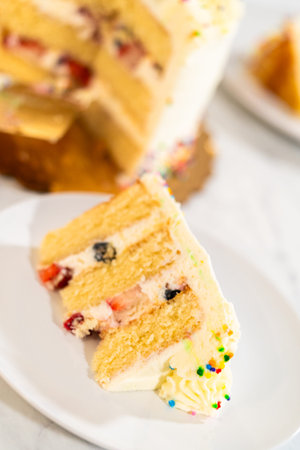 A vertical image of a slice of layered vanilla cake with colorful sprinkles and fruit filling on a...