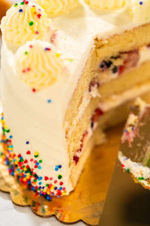 An angled close-up of a birthday cake with several piped frosting peaks and rainbow sprinkles along...