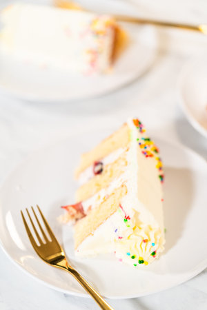 A vertical view of a slice of cake with smooth white frosting and pastel-colored sprinkles sits on a...