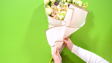 Hands present a neatly wrapped bouquet made with white roses, purple, and peach flowers. The wrap is beige with gold trim over a green surface.の写真素材
