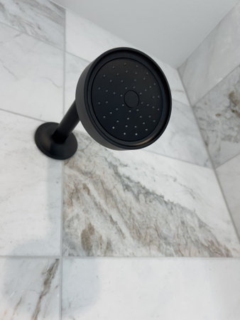View of a matte black shower head mounted on a veined marble-tiled wall. The shower head contrasts beautifully with the clean tile design. Perfect for home improvement content or interior design portfolios.の写真素材