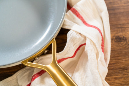 Two ceramic frying pans with golden handles resting on a red-striped dish towel over a wooden table. The light highlights the smooth surface of the cookware.の写真素材