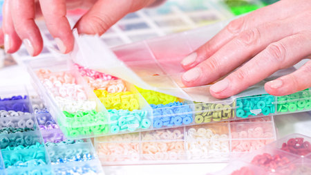 Woman hands gracefully poised over a collection of beads, sorted by color in transparent organizers....