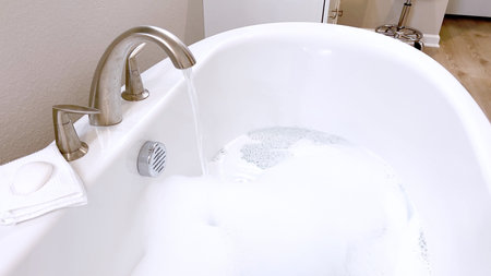 Elegant white bathtub filled with water, featuring a sleek brushed nickel faucet with water flowing...