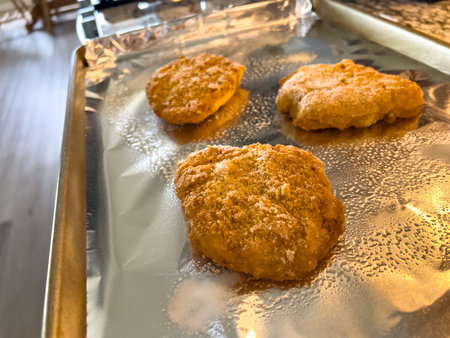 Three breaded chicken patties rest on an aluminum foil-lined tray on a dark kitchen counter. The...