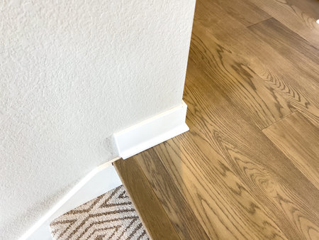 A view of a corner baseboard transition between two walls and wood flooring. The white trim and...