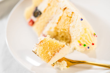 A detailed close-up of a golden fork lifting a bite-sized piece from a layered cake slice topped...