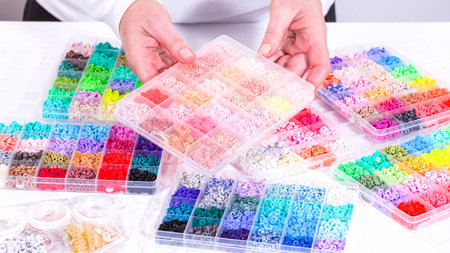 Woman hands gracefully poised over a collection of beads, sorted by color in transparent organizers....
