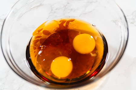 Close-up top view of a glass bowl containing two raw eggs and maple syrup, forming a dark base...