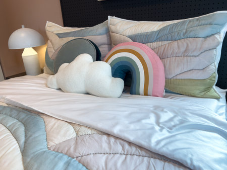 A cozy bed is decorated with rainbow and cloud shaped pillows. The room features modern lamps and a comfortable, cheerful interior design style.の写真素材