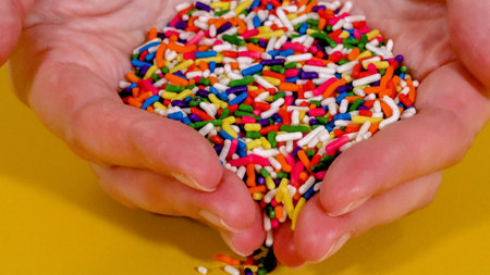 A close-up view shows hands holding a generous portion of rainbow-colored sprinkles. The vivid colors stand out on the yellow background.の写真素材
