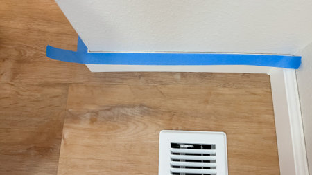 Blue painter tape is neatly applied along baseboards and a corner wall junction above a wood floor...