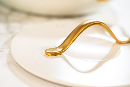 Macro shot of a sculpted golden handle resting on a white pot lid, highlighting luxury kitchenware elements. Perfect for product closeups and digital ads.の写真素材