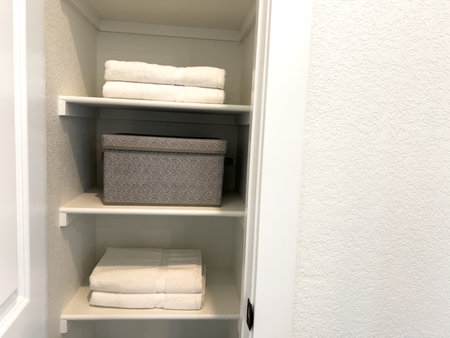 An organized linen closet with neatly folded white towels and grey fabric bins on white shelves. The space is built into a white interior wall and kept minimal.の写真素材