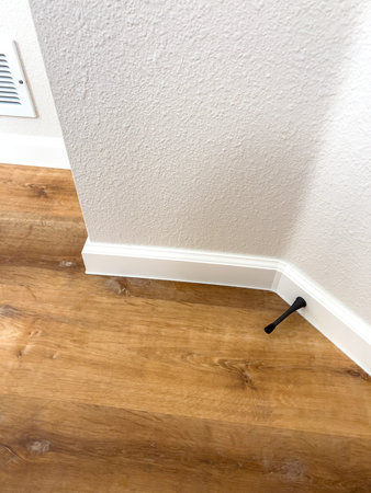 A close view of an interior corner shows wooden flooring, white baseboard, and a black pipe coming...