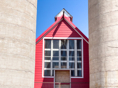 The facade of a red barn framed between two tall concrete silos. The barn has large windows and a peaked roof under clear daylight.の写真素材