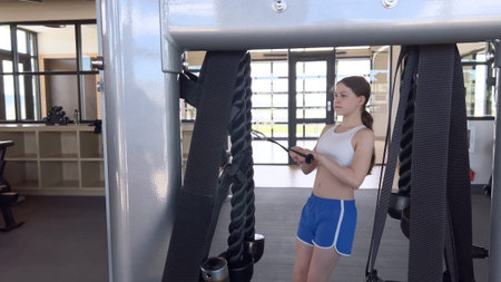 A young woman in athletic wear stands beside a gym cable machine in a bright workout space. The...