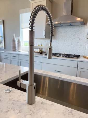 A tall stainless steel kitchen faucet with coiled pull-down hose sits above a large sink in a bright modern kitchen. The space features white cabinetry and light countertops.の写真素材