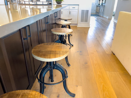 A row of industrial stools with wooden tops is set against a dark kitchen island on light hardwood flooring. The image highlights contrast and clean layout.の写真素材