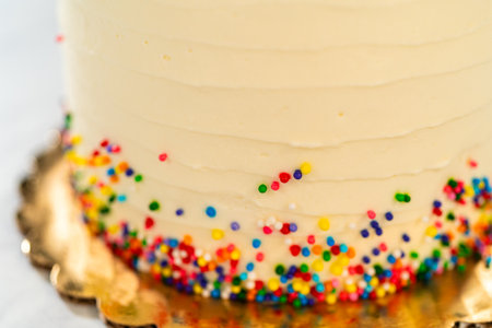 A close-up view of a cakes bottom edge featuring vibrant multicolored sprinkles against white frosting. The golden base board is partially in view. The focus is sharp on texture and sprinkle distribution.の写真素材