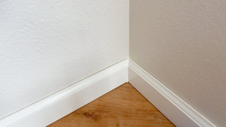 Close-up image shows a neat corner joint of white baseboards against a wood laminate floor and beige...