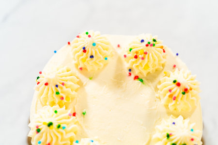 A close top-down view of a frosted white cake with piped rosettes made of whipped cream and decorated with rainbow sprinkles. The cake is placed on a white surface with natural light highlighting the textures.の写真素材