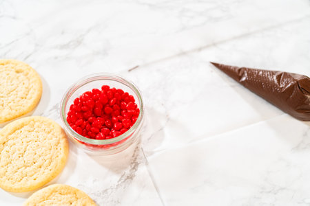 Top-down view of sugar cookies stacked in a black tray, alongside a piping bag filled with melted chocolate and a bowl of red candies, ready for decorating Easy Last Minute Reindeer Sugar Cookies. These simple ingredients transform plain cookies into festive holiday treats.の写真素材