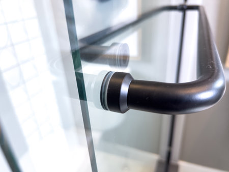 Close-up of a black shower door handle attached to a glass panel. The handle has a matte finish, and the image captures the transparency and reflection of the glass in bright lighting.の写真素材
