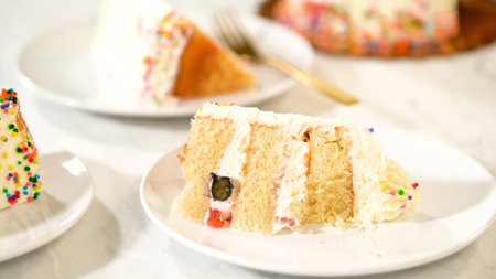 A slice of birthday cake with fruit and cream filling is displayed on a white plate. The cake is decorated with sprinkles and rests on a marble surface.の写真素材