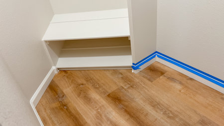 Wide-angle shot of closet baseboards masked with blue painter tape in a room with white walls and light wood flooring. The space appears ready for a painting update.の写真素材