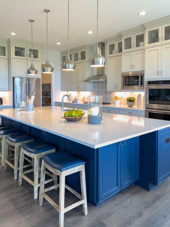 A large bright kitchen features a navy blue island with white countertops and modern pendant lighting. White cabinets and stainless appliances complete the look.の写真素材